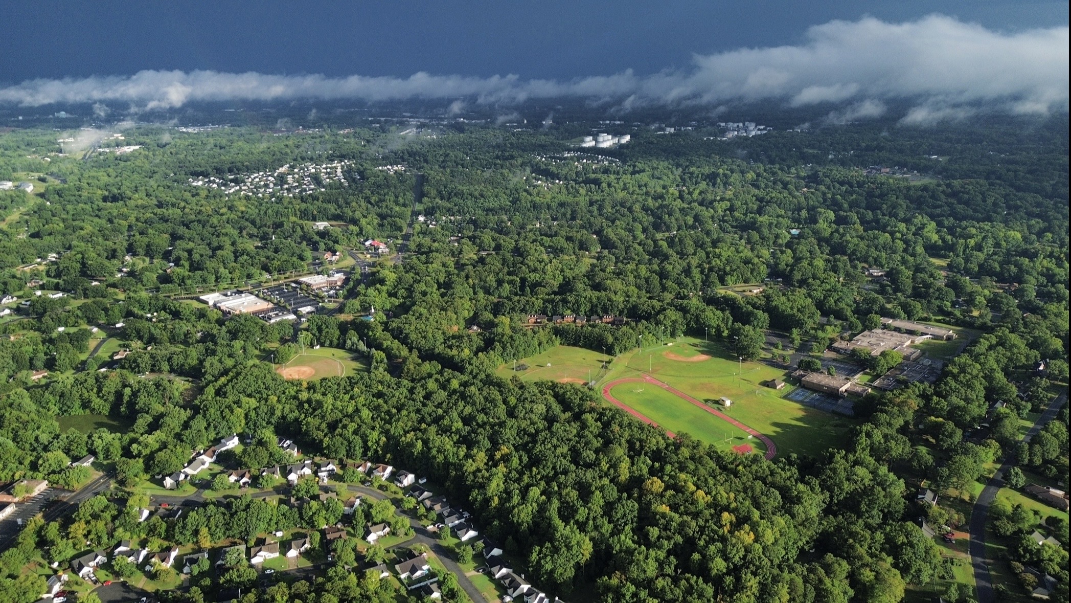 Daytime aerial view of athletic fields, surrounding properties, and a wooded neighborhood